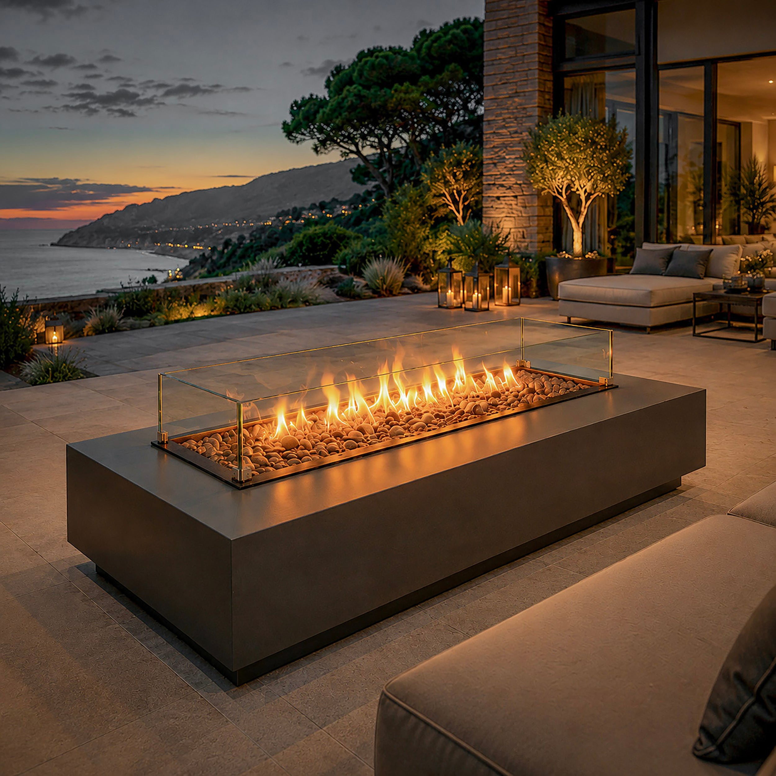 Aurora Flame luxe gas vuurtafel met glazen windscherm op een modern terras met uitzicht op zee tijdens zonsondergang.